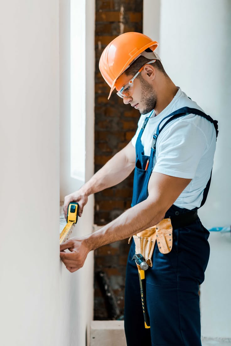 Professional worker measuring wall with tape measure, wearing orange hard hat and blue overalls