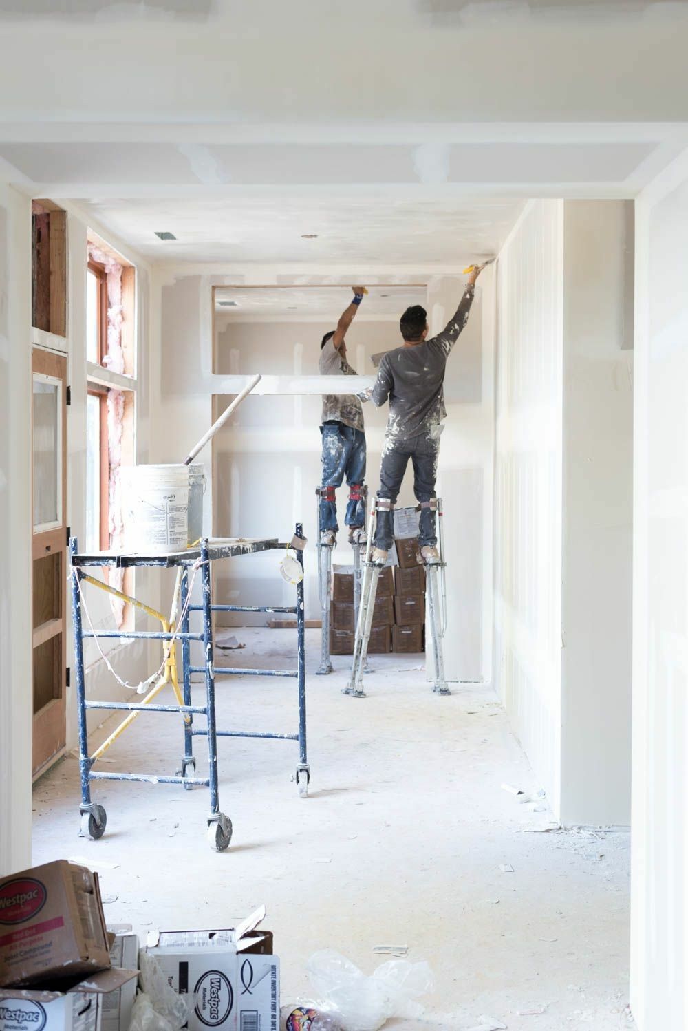 Two workers on stilts performing drywall finishing and painting in a bright room