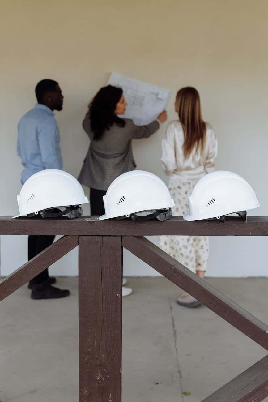Three white hard helmets on wooden beam, team reviewing blueprints in background